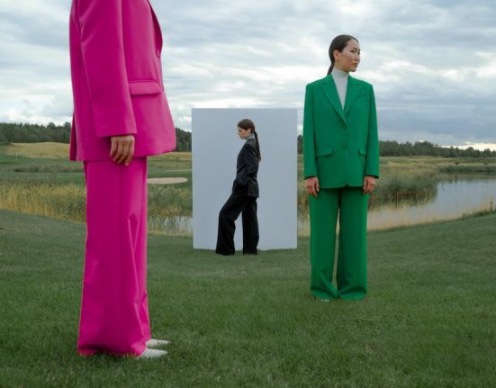 Models in colorful suits pose creatively on a grassy field beside a lake under a dramatic sky.