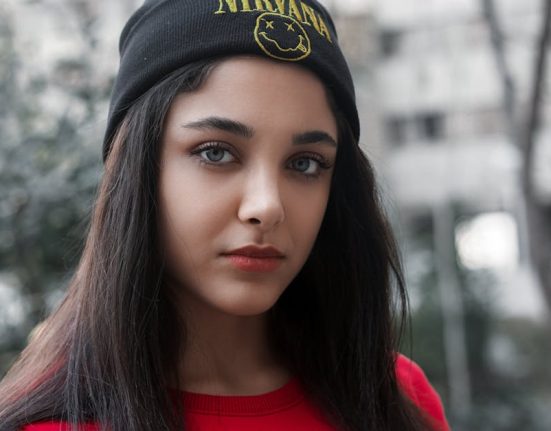 Close-up of a woman in a black beanie and red top with blurred urban background.